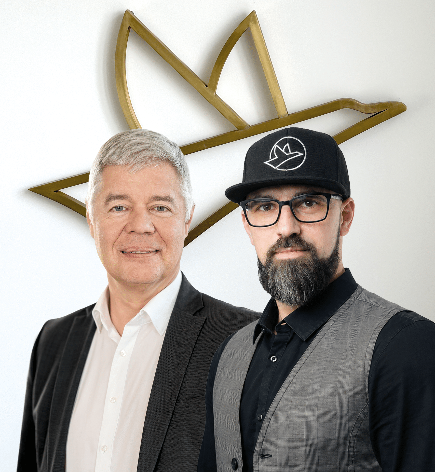 Zwei Männer stehen vor einer weißen Wand mit einem goldfarbenen Vogel-Logo, der linke trägt Anzug und weißes Hemd, der rechte trägt Brille, schwarzen Hut mit weißem Vogel-Logo, schwarzen Hemd und graue Weste