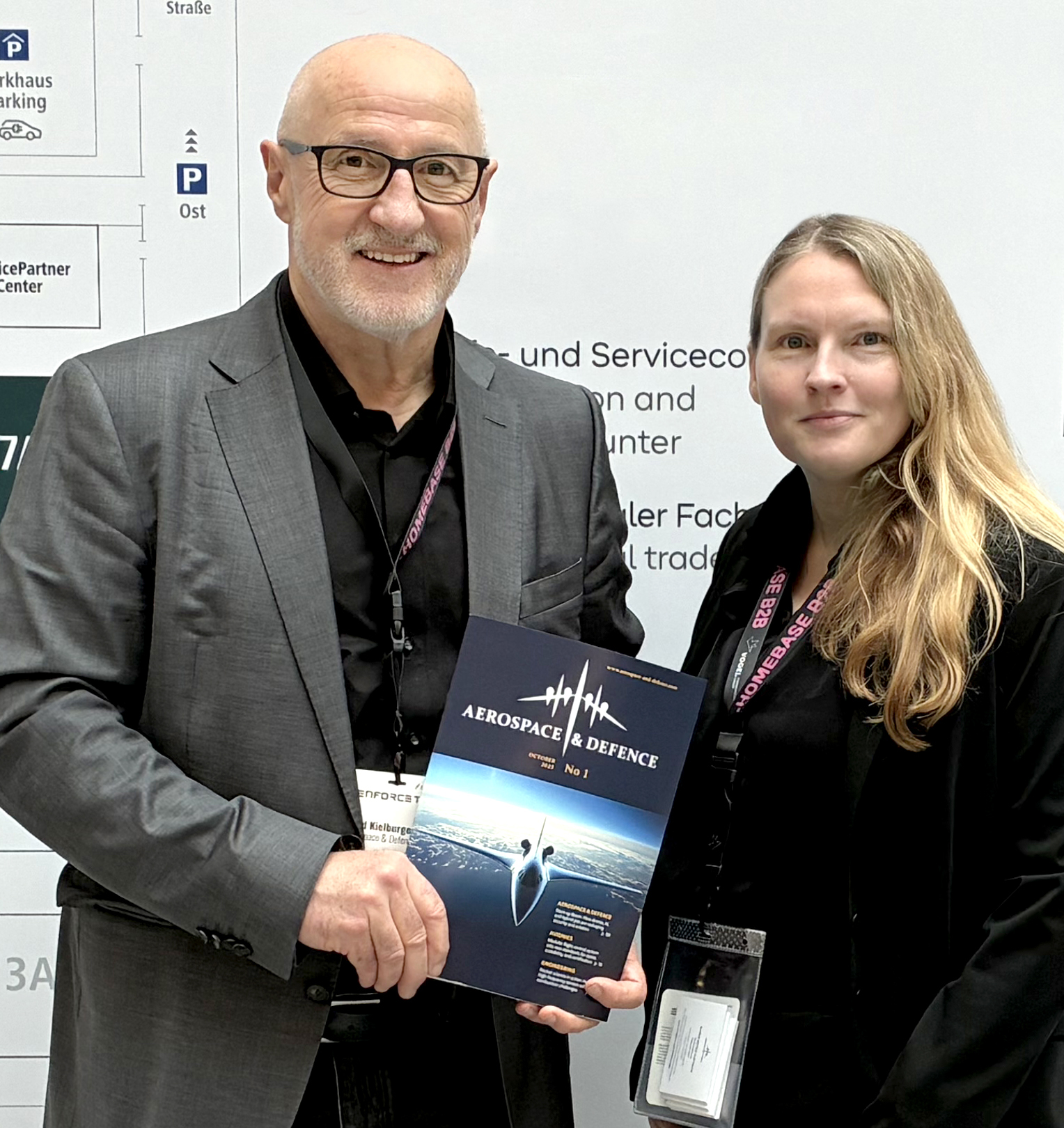 Zwei Personen in schwarzer Kleidung mit Namensschildern, der Mann hält eine Broschüre mit der Aufschrift 'Aerospace & Defence'
