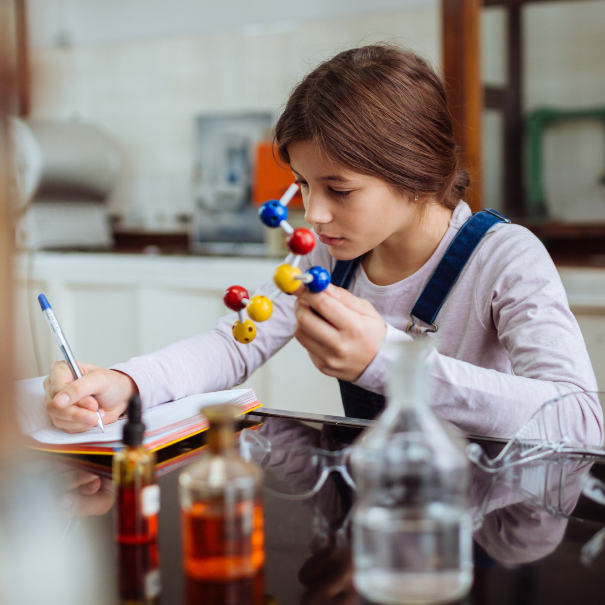 Mädchen hält Molekülmodell und schreibt in Notizbuch an einem Tisch mit Laborflaschen