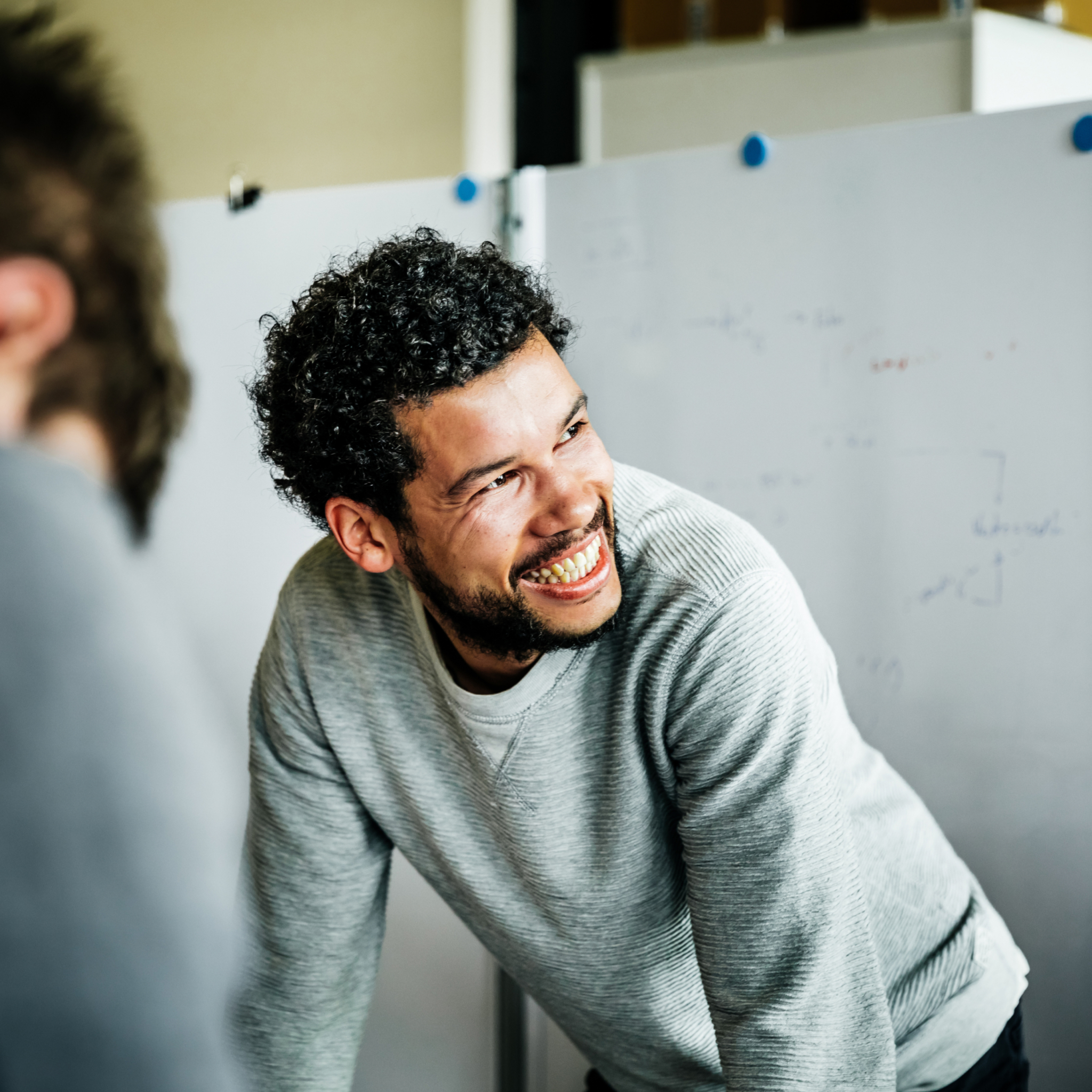 Lächelnder Mann mit lockigem Haar und grauem Pullover lehnt sich nach vorne in einem Büro mit Whiteboard im Hintergrund