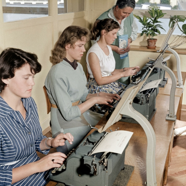 Drei junge Frauen sitzen nebeneinander an historischen Schreibmaschinen und tippen in einem hellen Raum mit Fenster und Pflanzen.